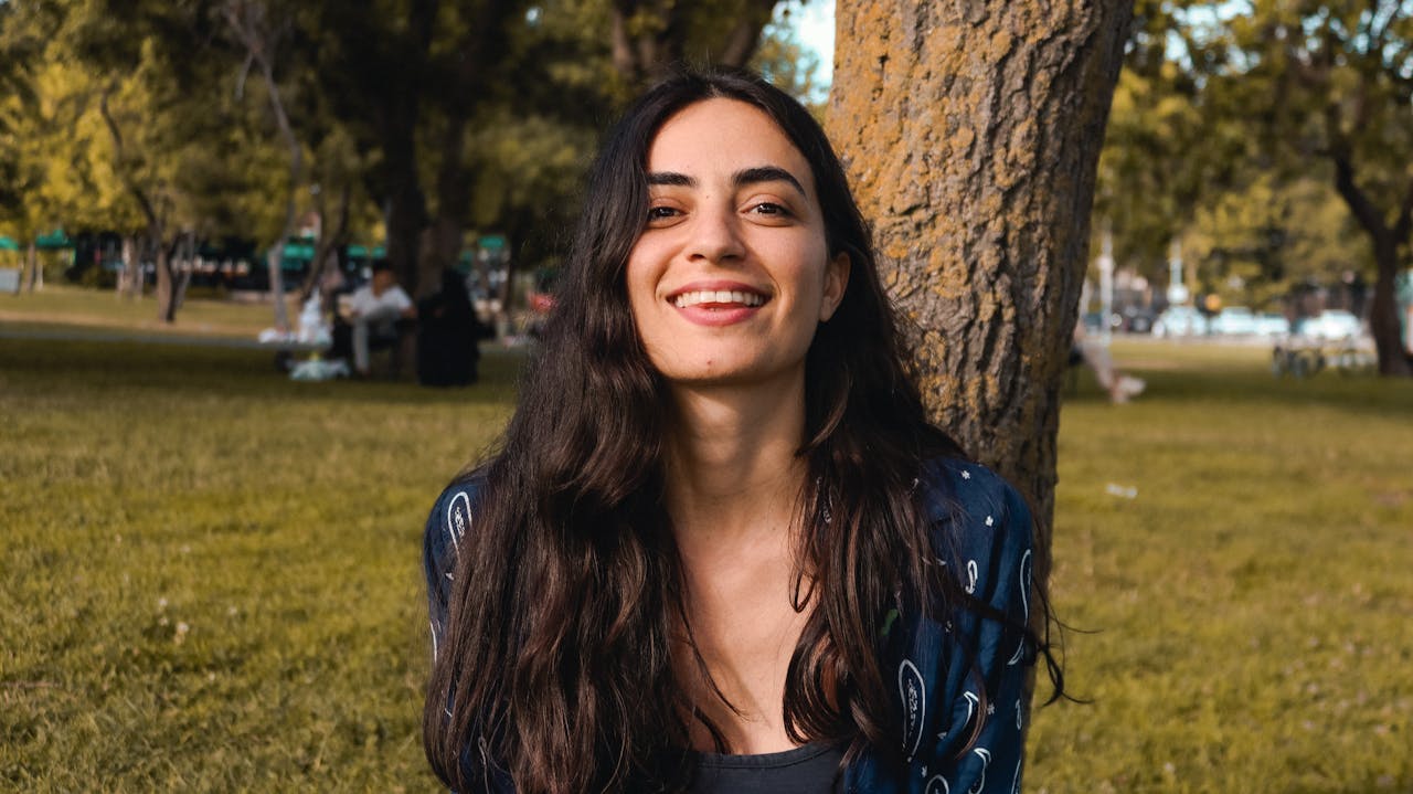 my-qualifications Smiling young woman leaning against a tree in a sunlit park.