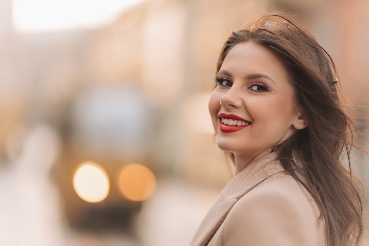 about-img Portrait of a smiling woman with red lipstick and brown hair outdoors.